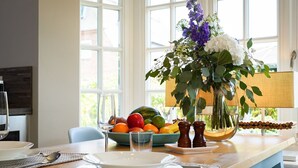 Een eettafel met borden, bestek, een fruitschaal, een vaas met bloemen en een raam met uitzicht.