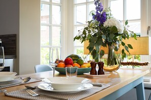 Ein Esstisch mit Tellern, Besteck, einer Obstschale und einer Vase mit Blumen, sowie ein Fenster mit Blick.