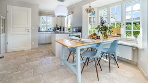Een moderne keuken met een centraal eiland, een zithoek met blauwe stoelen, en grote ramen die voor veel daglicht zorgen.