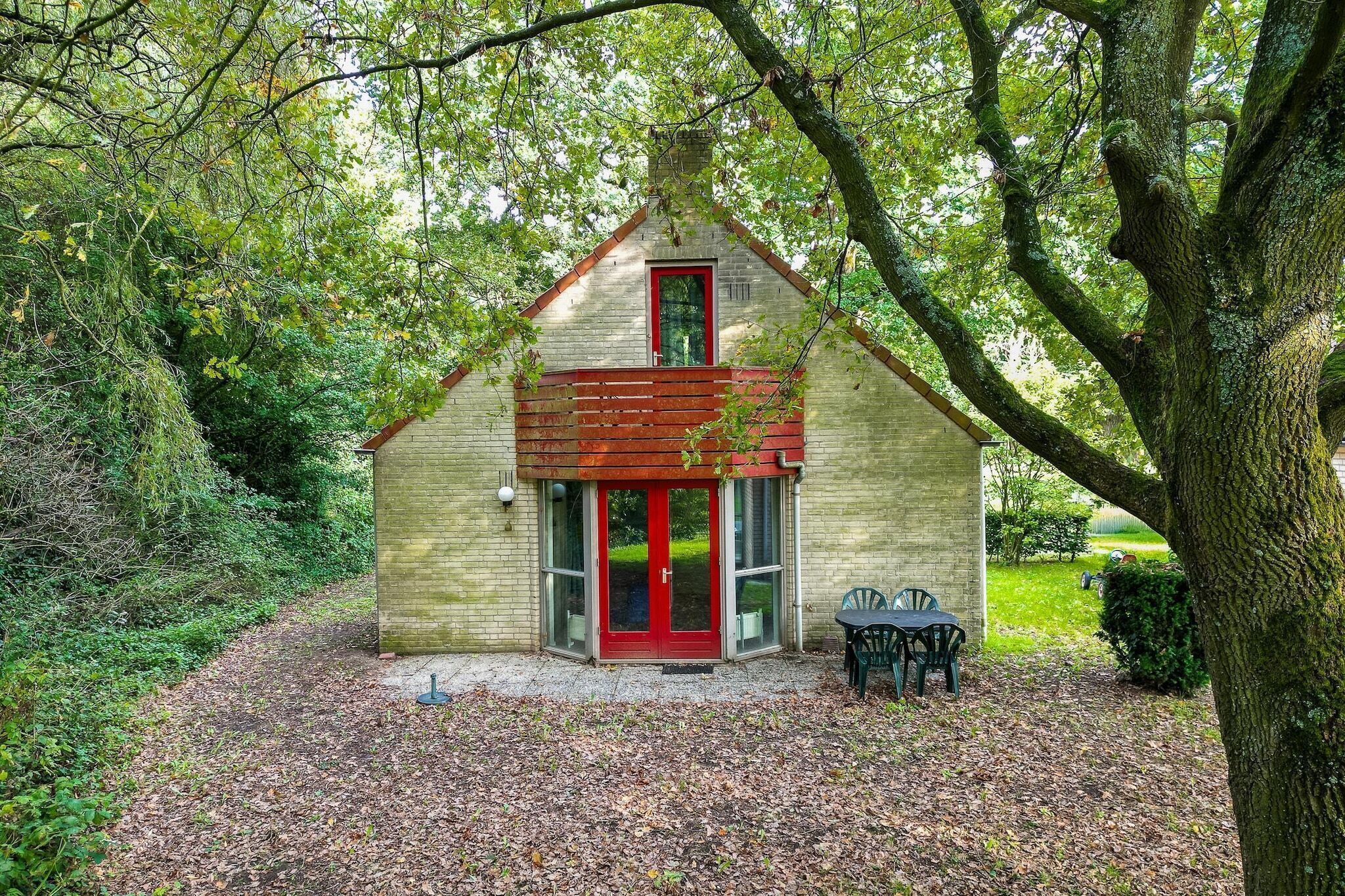 A small, single-story building with a red door and a brick exterior, surrounded by trees and a gravel path.