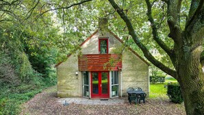 A small, single-story building with a red door and a brick exterior, surrounded by trees and a gravel path.