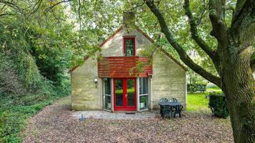 A small, single-story building with a red door and a brick exterior, surrounded by trees and a gravel path.