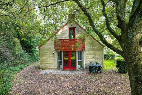 A small, single-story building with a red door and a brick exterior, surrounded by trees and a gravel path.