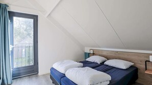 Une chambre avec un plafond en pente, une grande fenêtre avec des rideaux, un lit avec une literie blanche et bleue, et une tête de lit en bois.