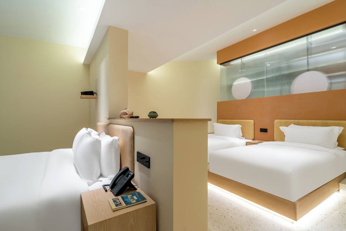 A hotel room with two beds, a bedside table with a phone and a book, a small shelf, and a large window with a view.