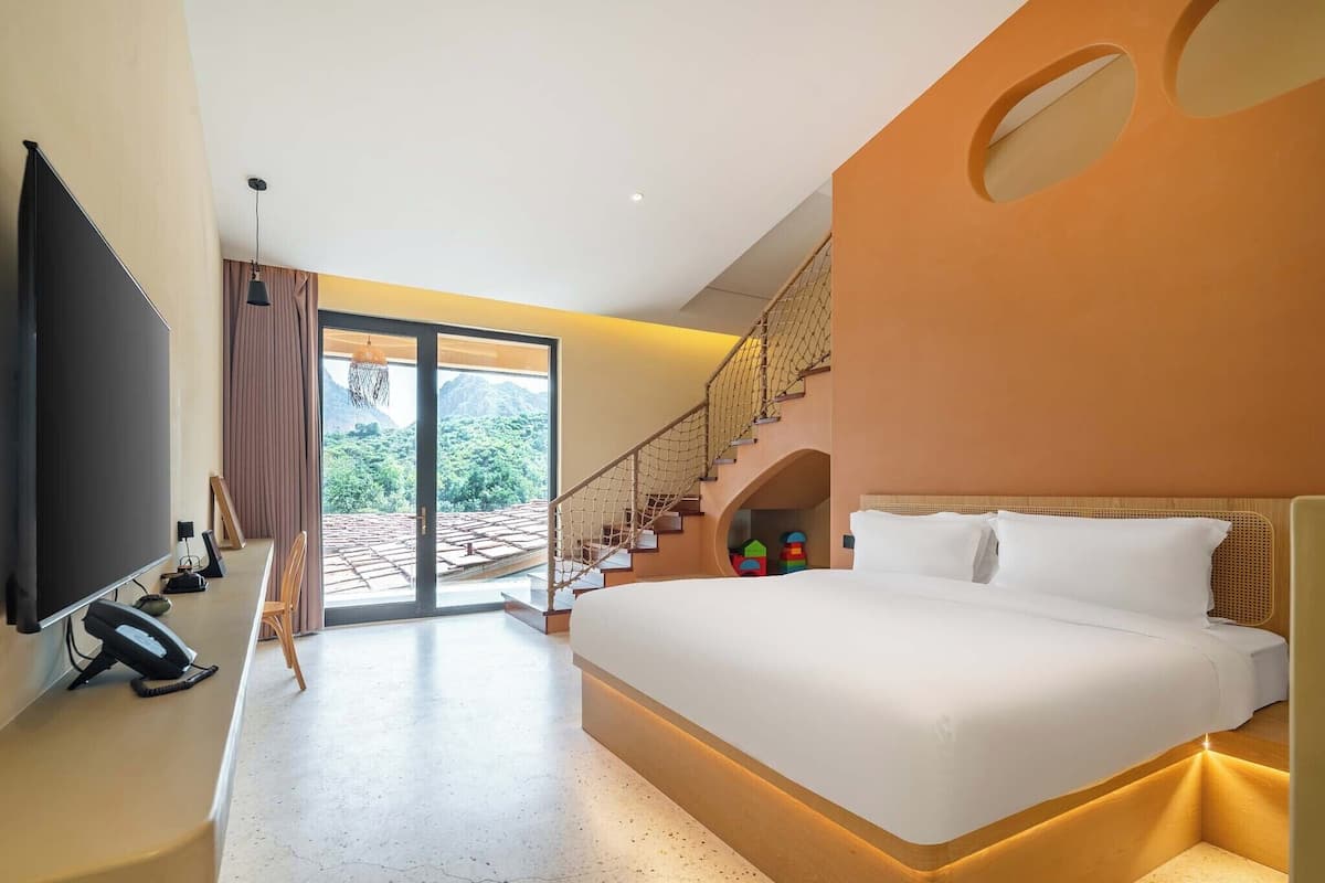 A modern hotel room with a large bed, a flat-screen TV, a desk with a telephone, and a staircase leading to a balcony with a mountain view.