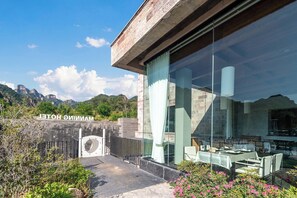 A modern hotel with a glass facade, outdoor seating, and a mountainous backdrop.