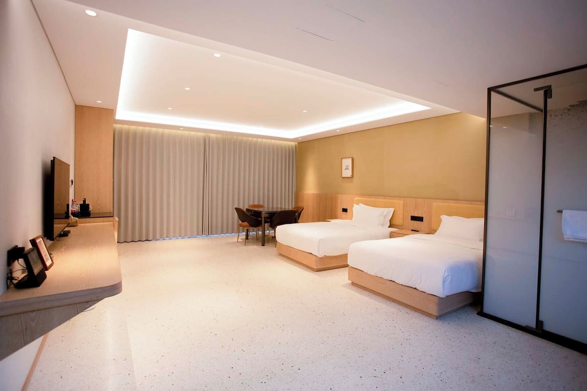 A hotel room with two beds, a desk, a chair, a TV, and a large window with curtains.