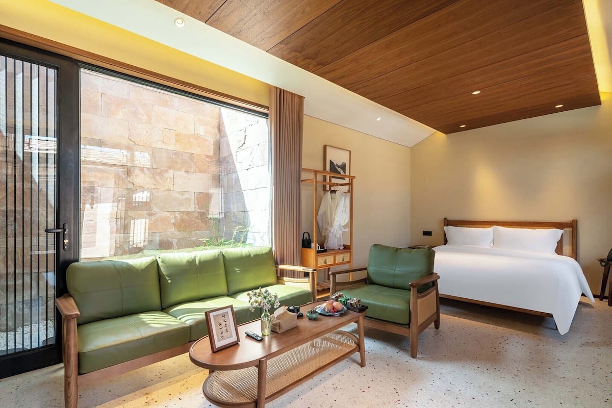 A hotel room with a bed, two green sofas, a wooden coffee table, a mirror, and a large window.