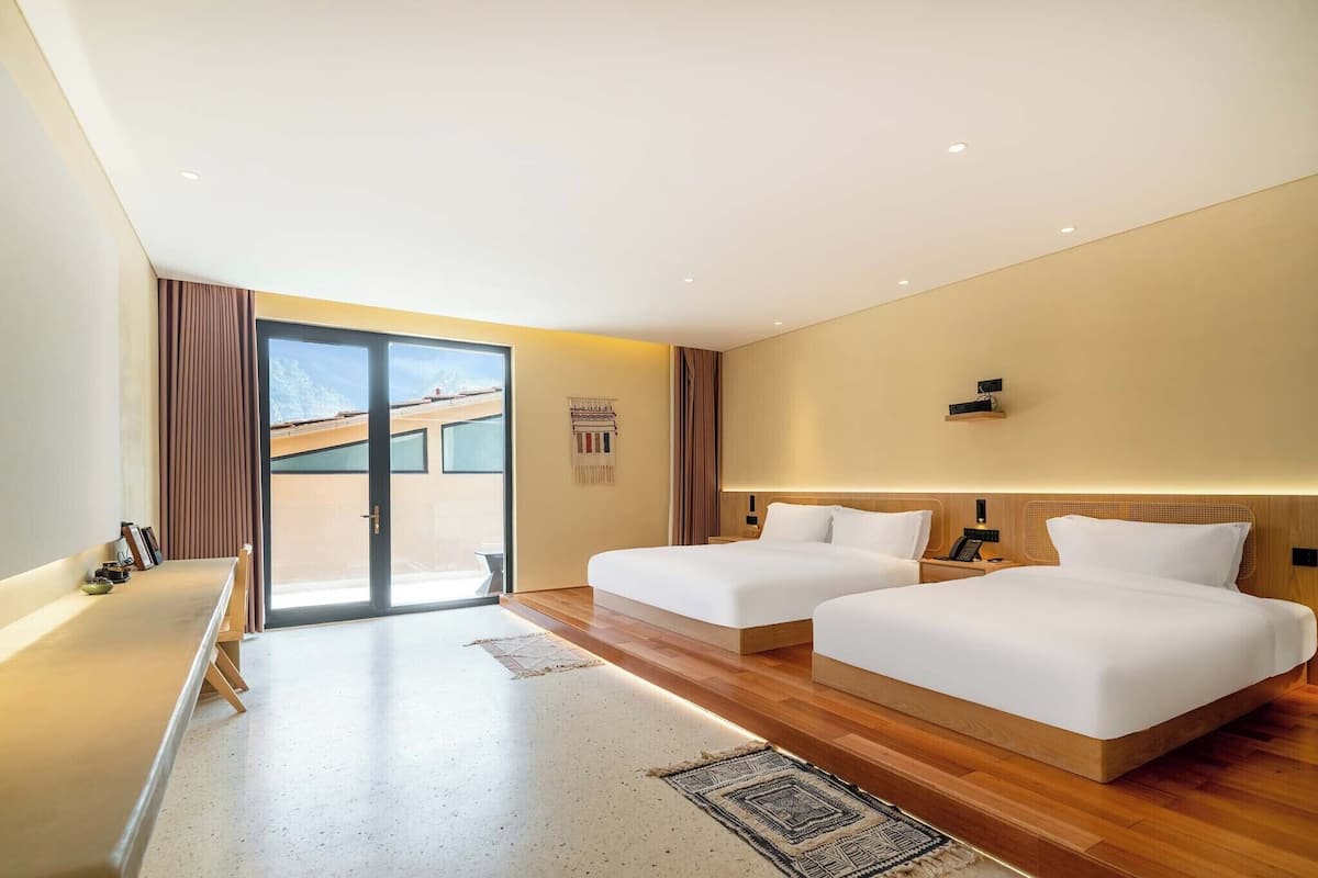 A modern hotel room with two beds, a large window, a desk, and a view of the outside.