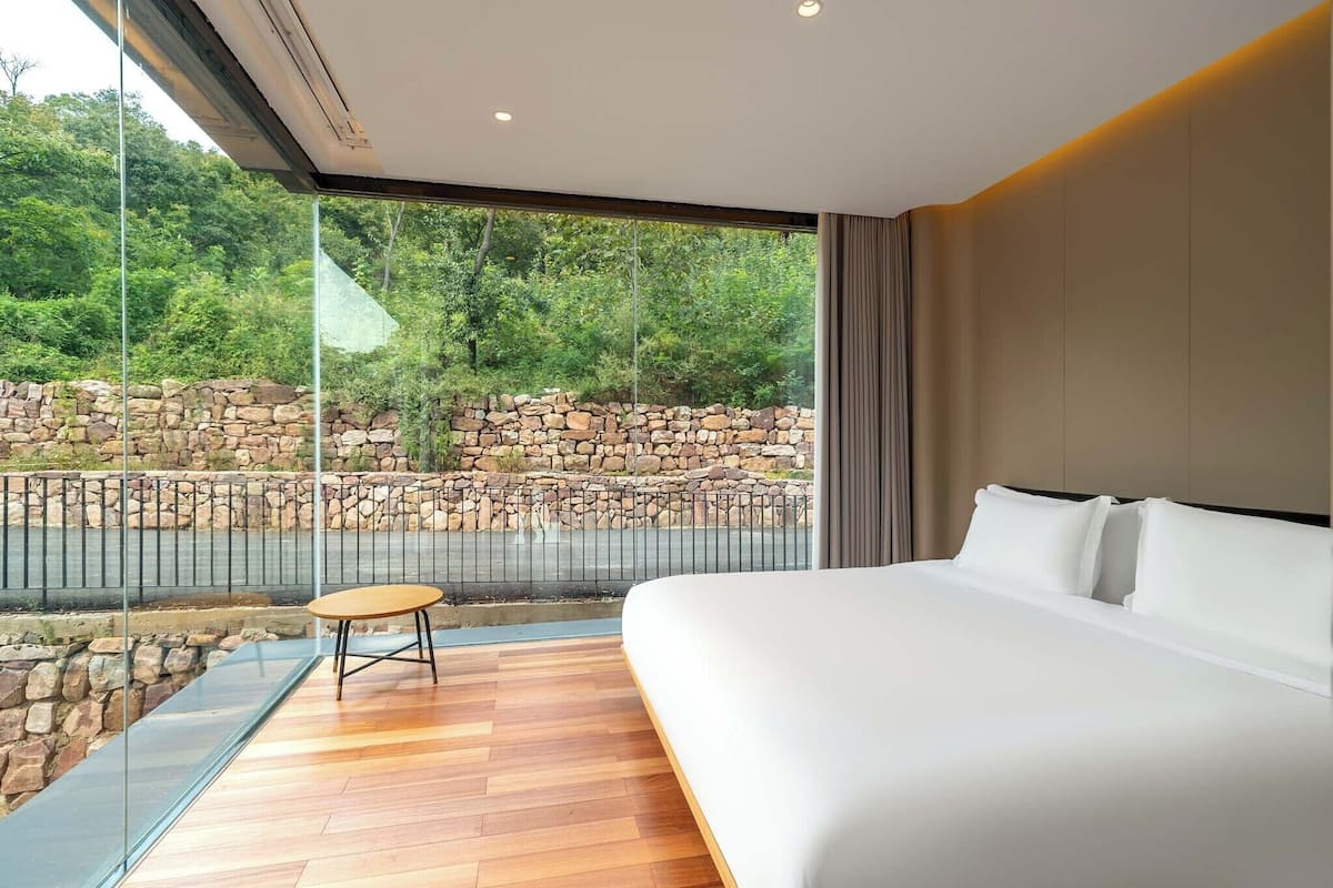 A modern hotel room with a large bed, a small table, and a view of a stone wall and greenery outside.