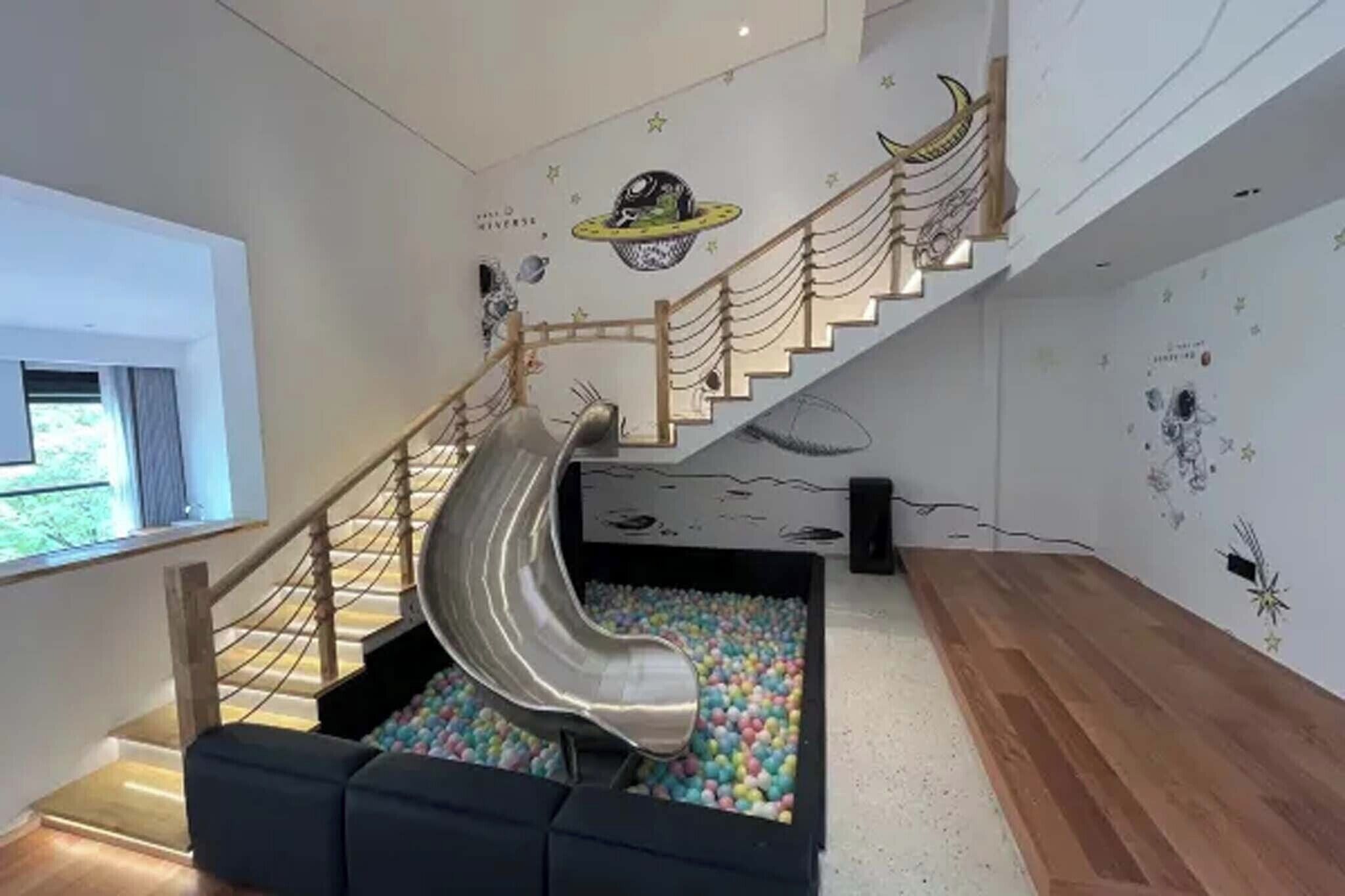A modern interior with a slide into a ball pit, a staircase with a wooden railing, and wall decorations.