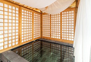 A traditional Japanese onsen with wooden lattice screens, a glass-enclosed hot spring, and a stone platform.