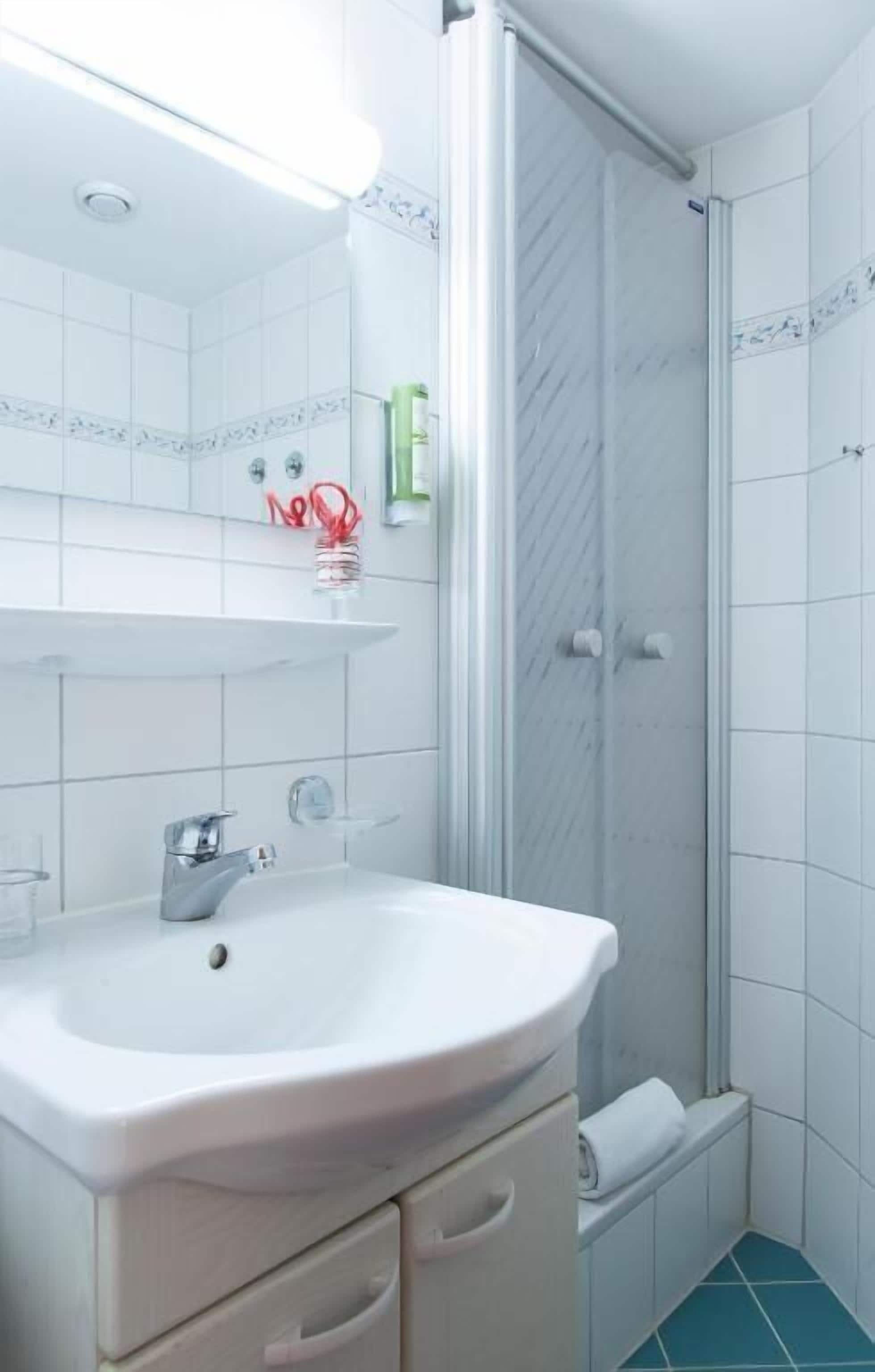 Un baño con lavamanos blanco, cabina de ducha de vidrio y suelo de baldosas.