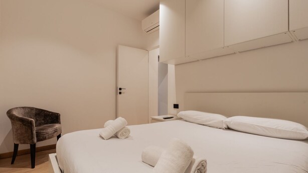 Hotellihuone, jossa on sänky, tuoli, yöpöytä ja ilmastointilaite.