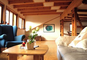 Une pièce avec un plafond en bois, un fauteuil bleu, un canapé beige, une table basse en bois avec une bougie et un vase rempli de fleurs.