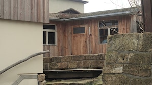 Eine Steintreppe führt zu einem Holzhaus mit einer Steinsäule und einer Bank, auf der eine Tasche und Schuhe liegen.