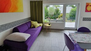 Ein Zimmer mit einem violetten Sofa, einem kleinen Tisch mit einer Lampe und einem Fenster mit Blick auf Grünflächen.