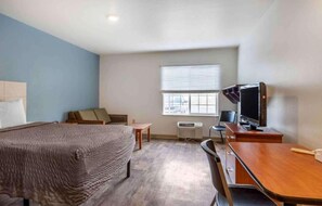 A hotel room with a bed, a desk with a computer, a chair, a window with blinds, and an air conditioning unit.