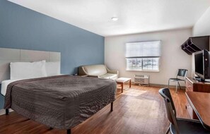 A hotel room with a bed, a desk with a computer, a chair, a window with blinds, and a television.