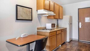 A compact kitchen with wooden cabinets, a microwave, a refrigerator, a small table with a chair, and a framed picture on the wall. - 1 Full Bed | Mobil Acc RI Shwr Nsmk Kitchen (Wichita KS)