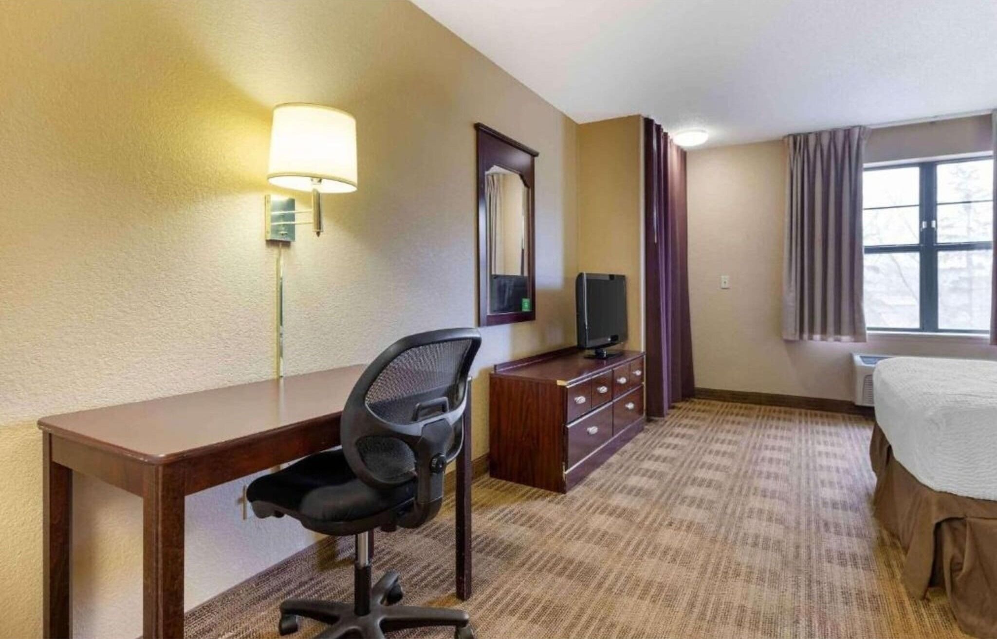 A hotel room with a desk, chair, TV, dresser, and a bed.
