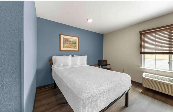 A single bed with white bedding in a room with blue walls, a framed picture on the wall, a window with blinds, and a wall-mounted heater.