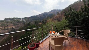 A balcony with wicker furniture and a table set for a meal, overlooking a hilly landscape. - Alaya Stays Amara Dawn (Naina Range)