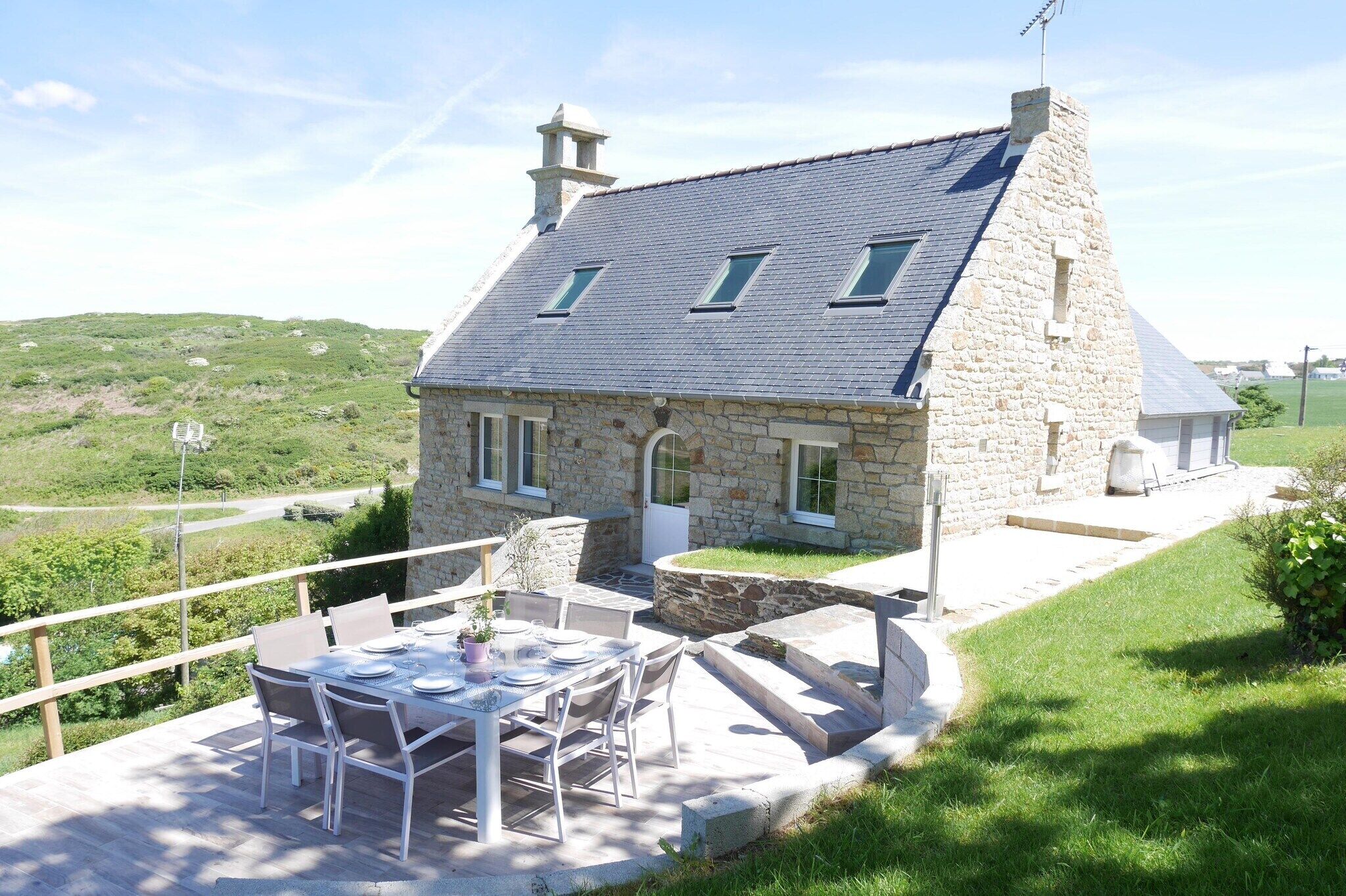 A stone house with a sloped roof, a balcony with a table and chairs, and a view of the surrounding landscape.