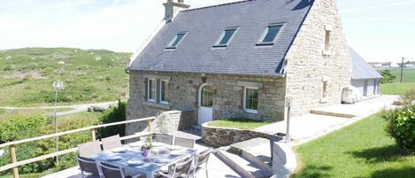 A stone house with a sloped roof, a balcony with a table and chairs, and a view of the surrounding landscape.