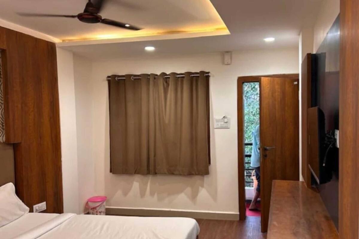 Un dormitorio con una cama, ventilador de techo, ventana con cortinas, banca de madera y televisor montado en la pared.