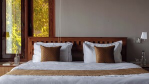 A neatly made double bed with white linens and two brown pillows, a wooden headboard, a bedside table with a lamp, and a window with a view of trees. - Alaya Stays Amara Twilight (Naina Range)
