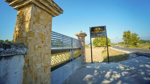 A stone gate with a metal fence, a sign for JAI GANESHA RESORT, and a road leading to a building with a dome. - Jai Ganesha Resort (Mal)