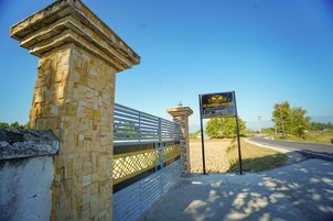A stone gate with a metal fence, a sign for JAI GANESHA RESORT, and a road leading to a building with a dome.