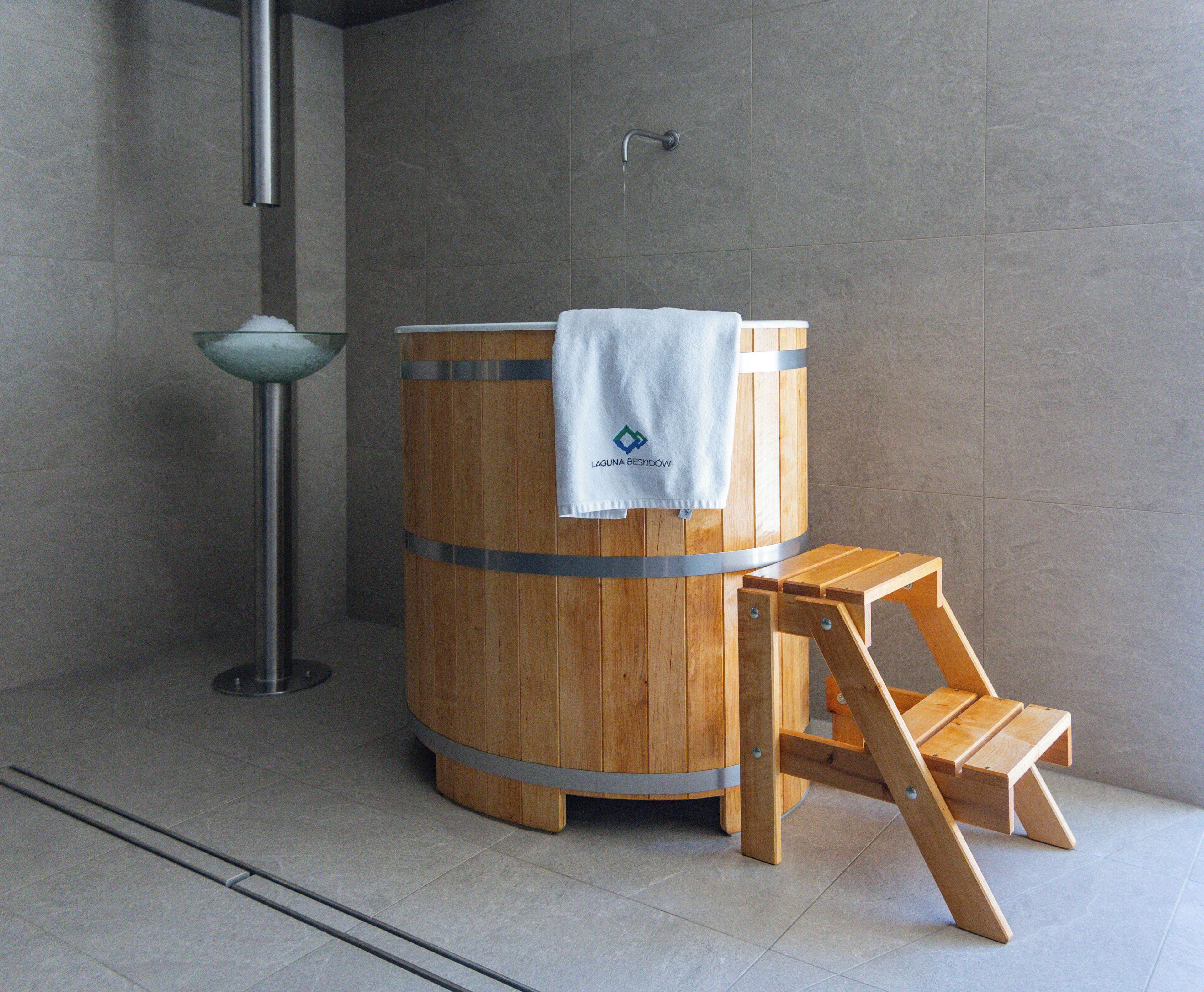 A wooden barrel-shaped sauna with a towel on top, a wooden stool, and a metal basin.