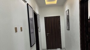A hallway with white walls, framed pictures, a wooden door, and a ceiling light fixture. - Aurora Smart Residence (Abuja)