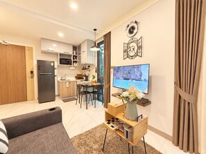 A modern living room with a sofa, a coffee table with a vase, a TV on the wall, and a kitchen in the background. - A103-  1 Bedroom Serviced Apartment with Whirlpool at Ao Nang Beach (Tambon Ao Nang)
