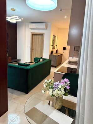 Une chambre d’hôtel moderne avec un canapé vert, une petite table ornée d’un vase de fleurs, un bureau avec une chaise et une salle de bain équipée d’un lavabo et d’un miroir.