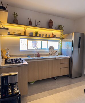A modern kitchen with a stainless steel refrigerator, a gas stove, a sink with a faucet, and a window above the sink. - Vrbo House - Praia do Francês AL (Marechal Deodoro)
