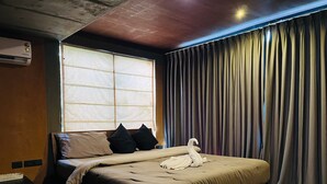 Quarto de hotel com cama, janela com persianas, cortina, ar condicionado fixado na parede e uma mesa pequena.