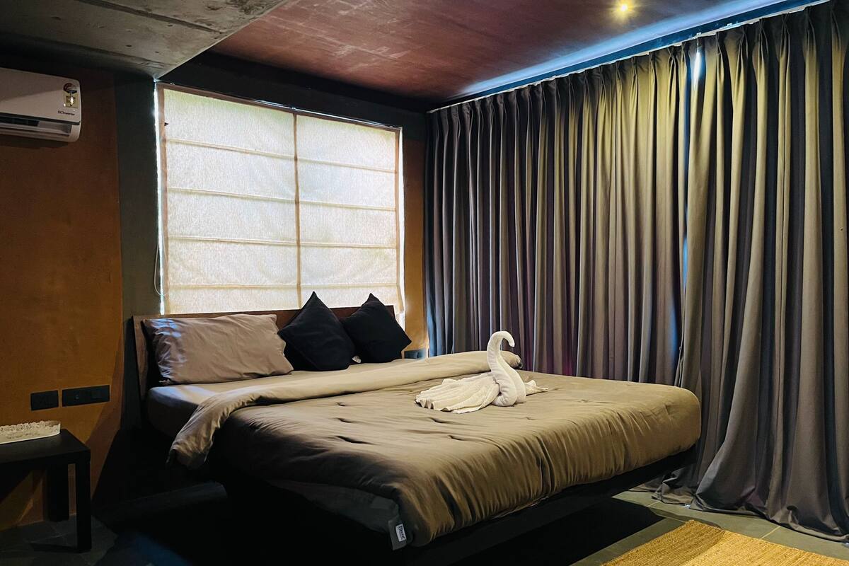 Una habitación de hotel con una cama, una ventana con persianas, una cortina, un aire acondicionado instalado en la pared y una pequeña mesa.