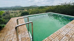 Piscina retangular com deck de madeira e guarda-corpos de metal, cercada por vegetação e montanhas ao fundo.