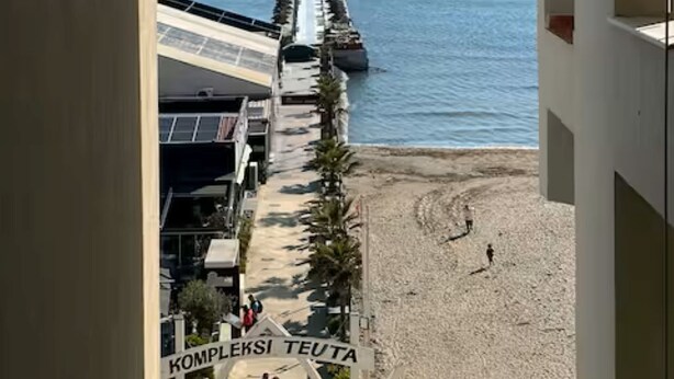 Blick auf einen Strand mit einem Steg, der ins Meer führt, einem Schild mit der Aufschrift 'KOMPLEKSI TEUTA' und einem von Palmen gesäumten Weg.