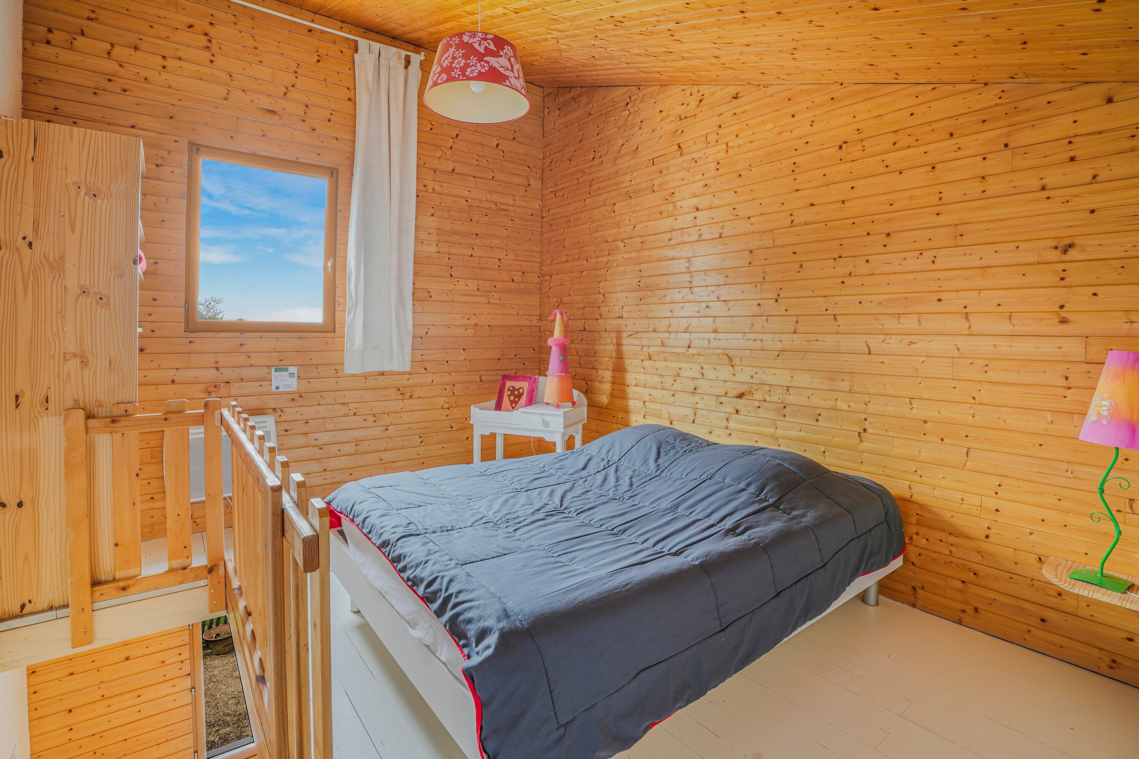 Quarto de cabana de madeira com beliche, uma mesinha com abajur e uma janela com vista.