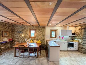 A rustic kitchen with stone walls, wooden beams, a dining table with chairs, and a built-in island with a sink.