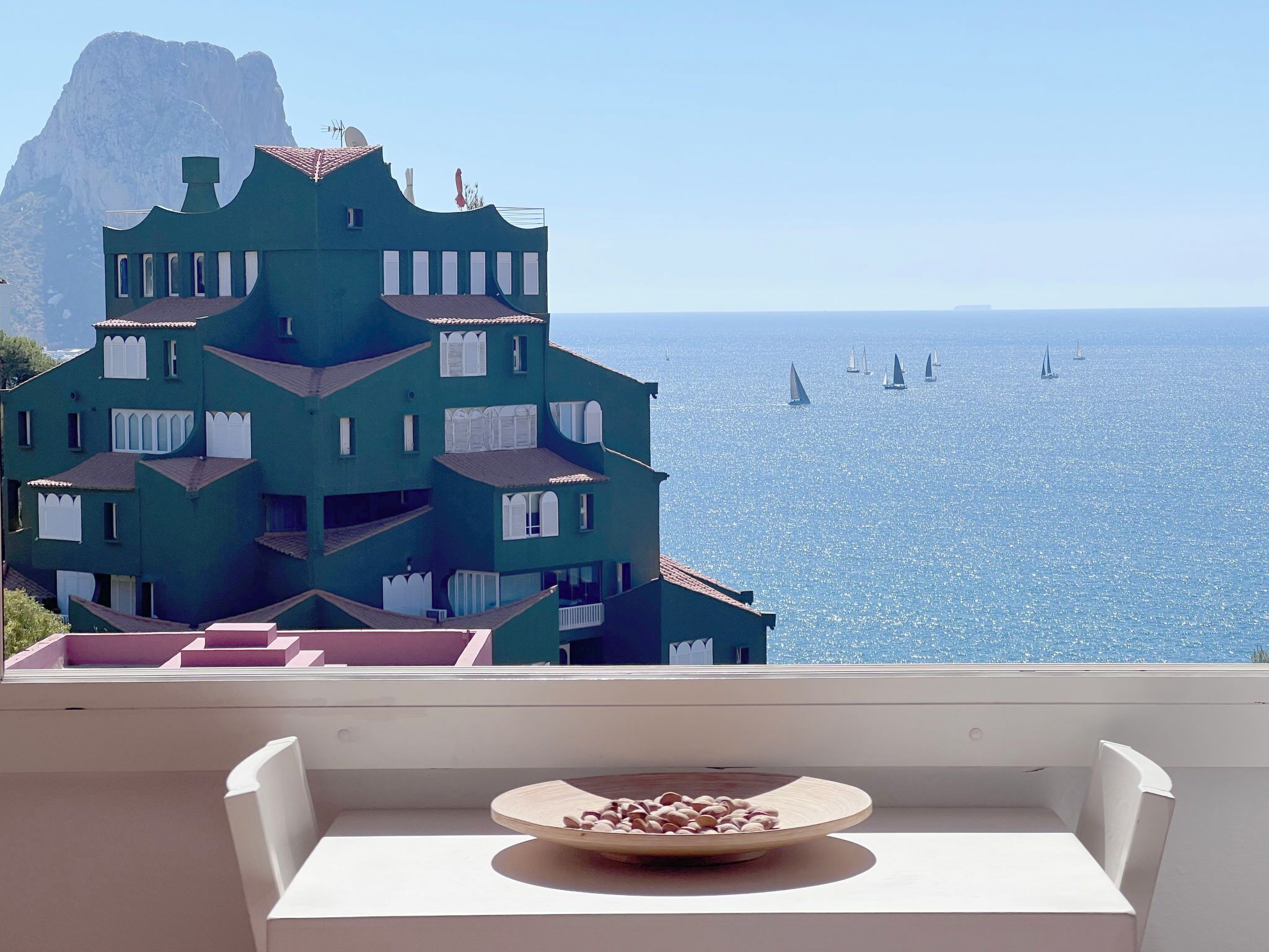 A balcony with a white table and a bowl of nuts, overlooking a sea with sailboats and a distinctive building with multiple balconies.