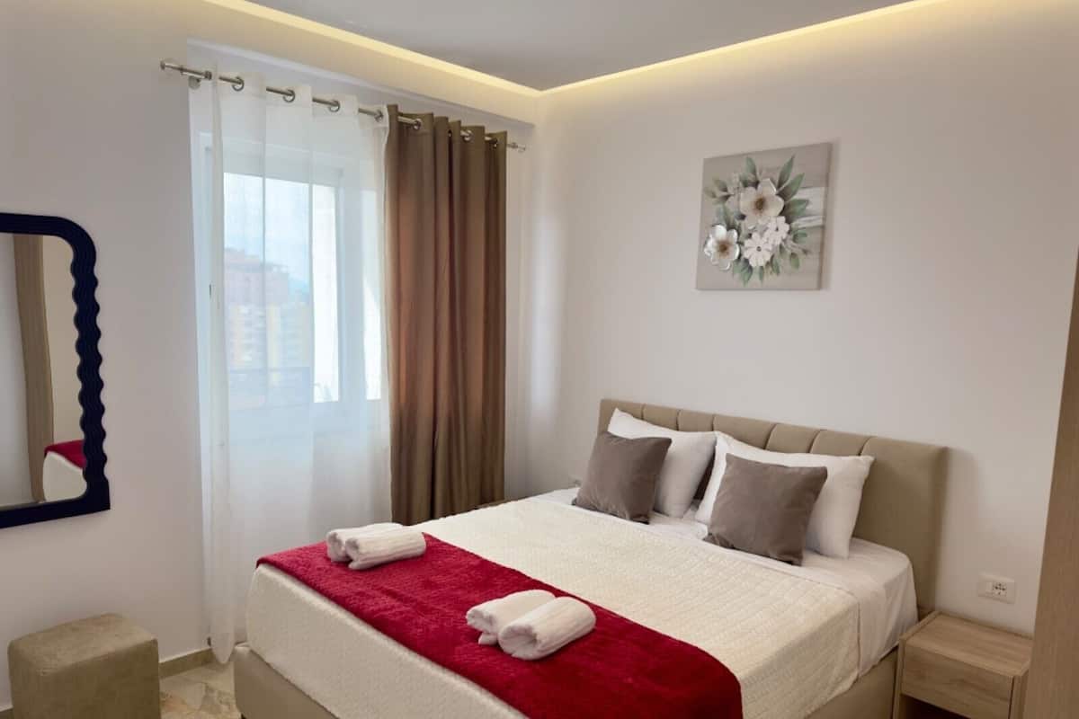Habitación de hotel con cama, mesita de noche, un taburete pequeño, espejo y ventana con cortinas.