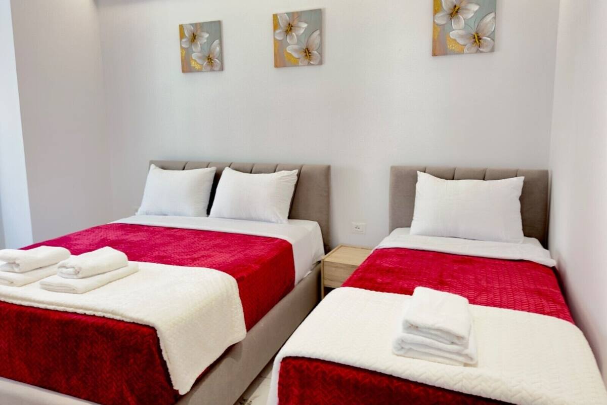Una habitación de hotel con dos camas, cada una con una colcha roja y blanca, almohadas blancas y una mesita de noche pequeña con una lámpara. Hay tres cuadros colgados en la pared sobre las camas.