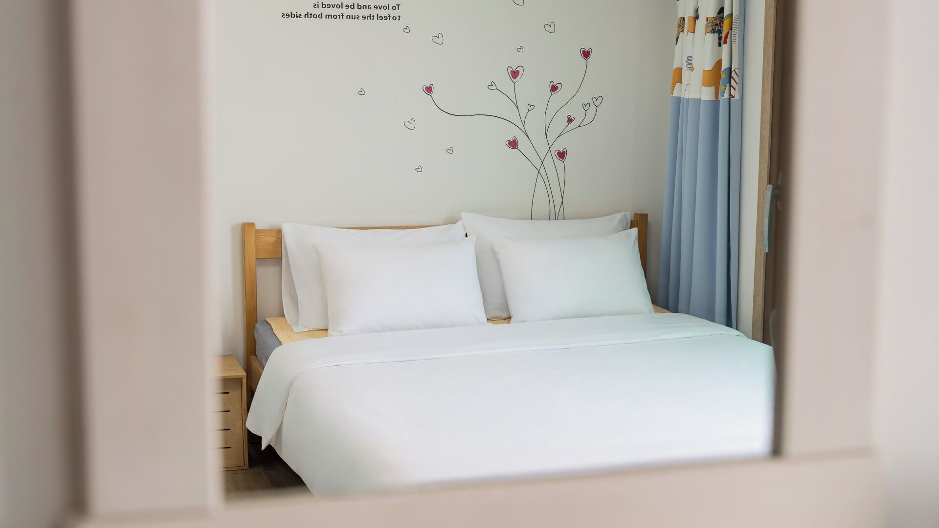 Una cama bien hecha con sábanas blancas, cabecera de madera y decoración de pared con un árbol y corazones.