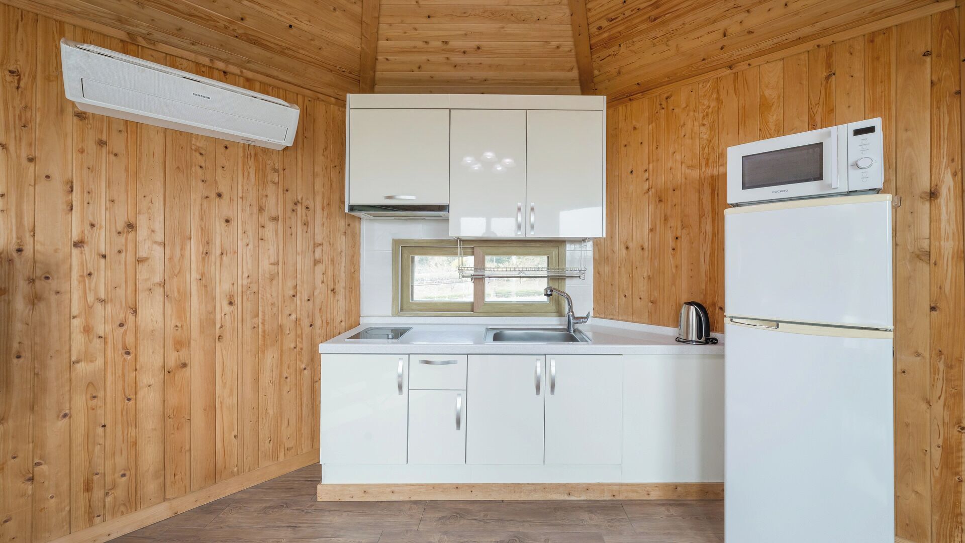 A compact kitchen with wooden walls, a window, white cabinets, a sink, a microwave, and a kettle.